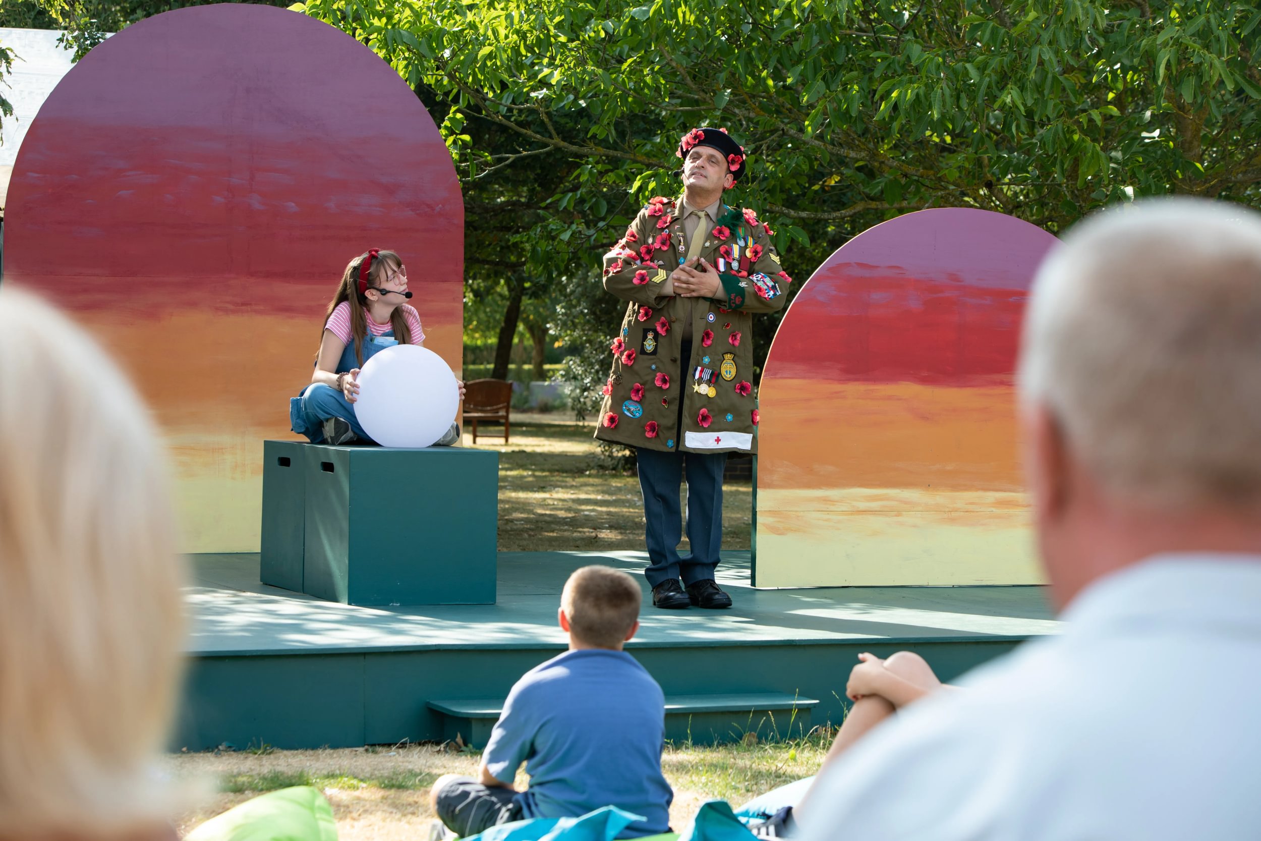 A young girl sits holding a glowing white orb. Next to her a man dressed in a large coat adorned with poppies, looks curiously down at her.