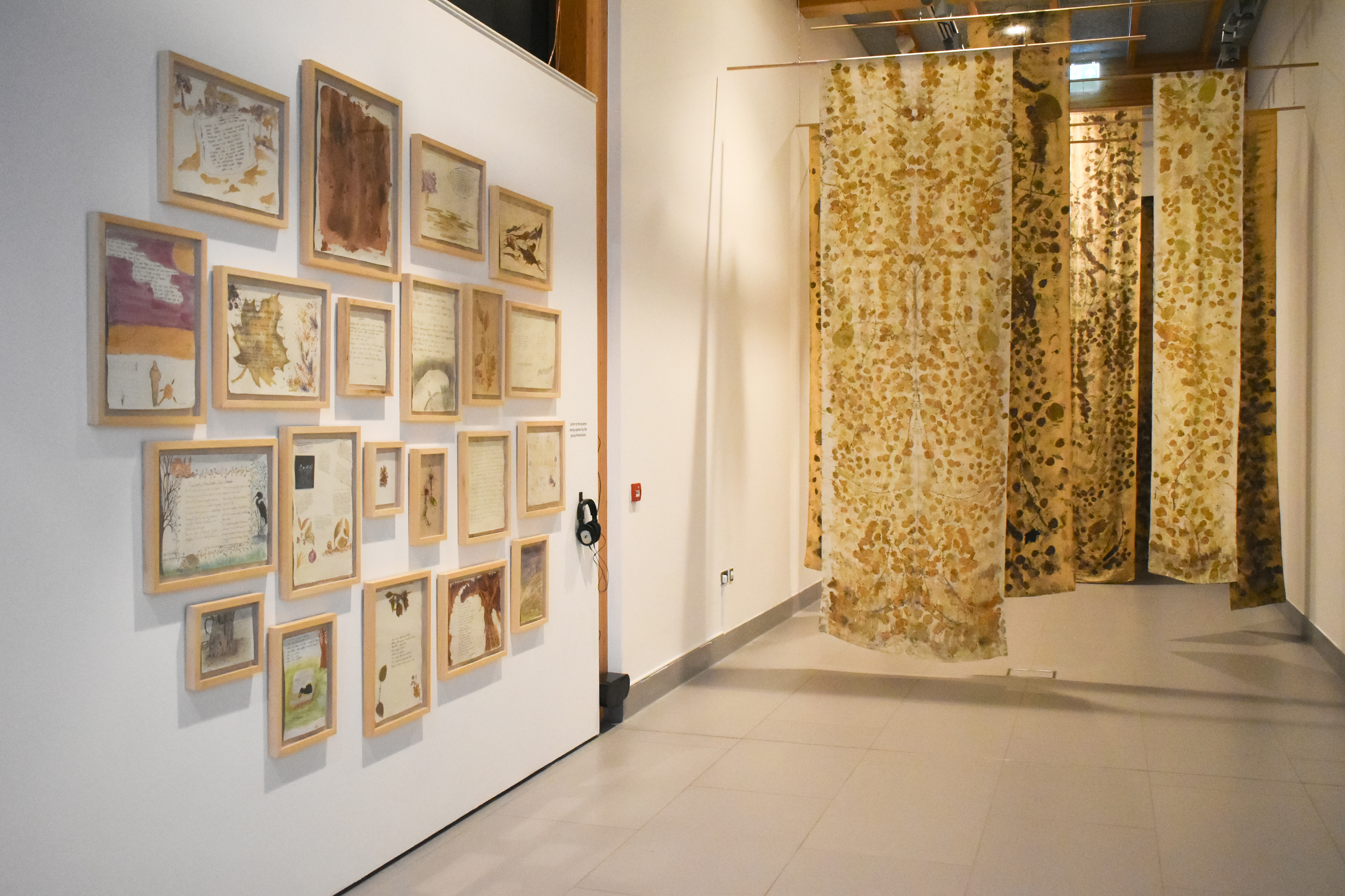 The Breaking Ground exhibition, part of Lockdown Landscapes. Framed artworks are hung on the wall, and silk, eco-dyed hangings stretch down from the ceiling.