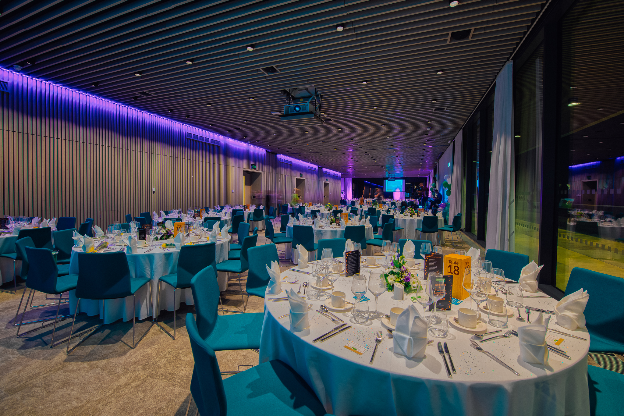 a large room is laid with tables and chairs, seemingly for a fancy dinner. It is night time as the sky is dark outside, and the lights are on.