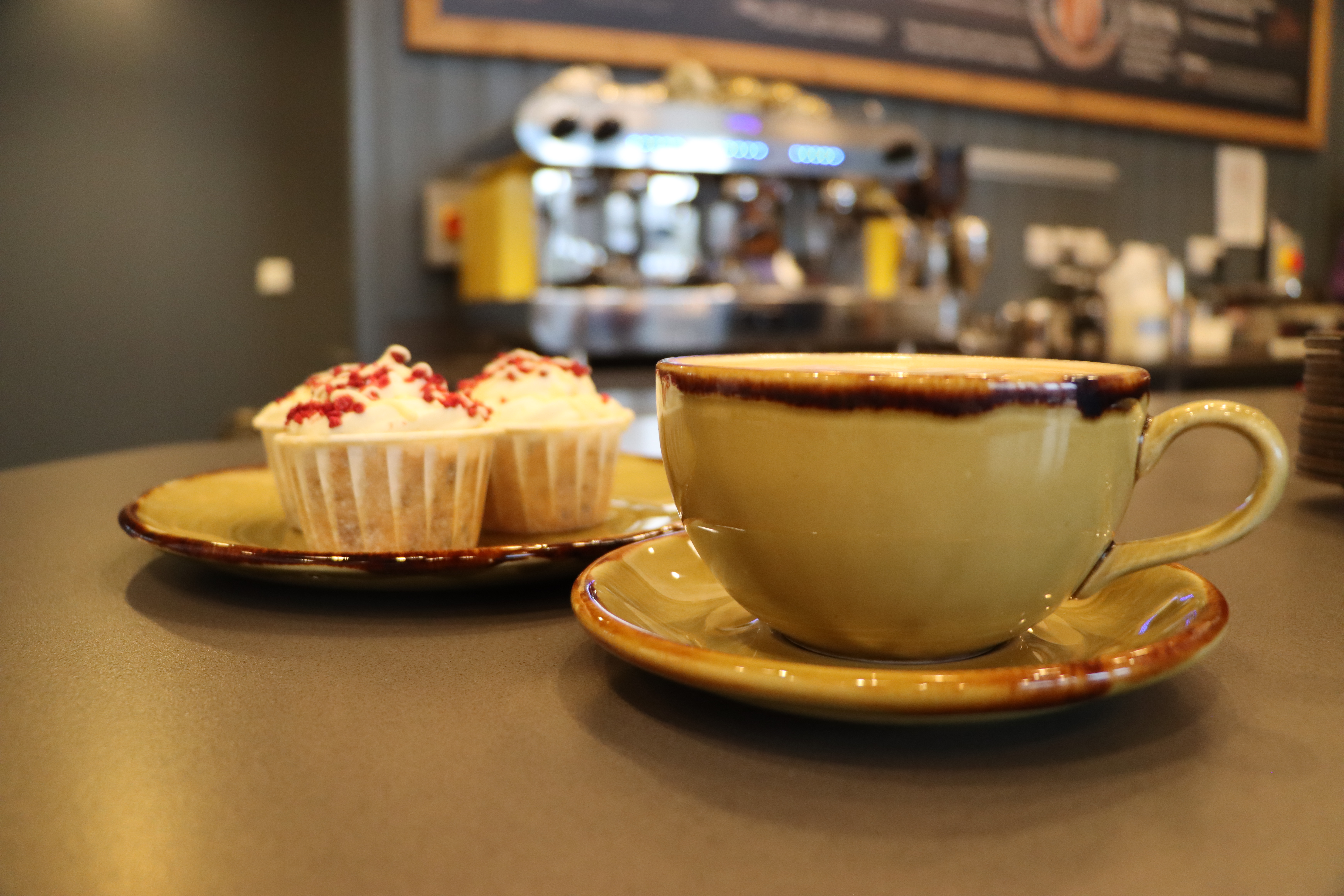 A cup of coffee in a large mug, sits in front of a plate of cupcakes within the National Remembrance Gardens coffee shop. The coffee machine is in the background.