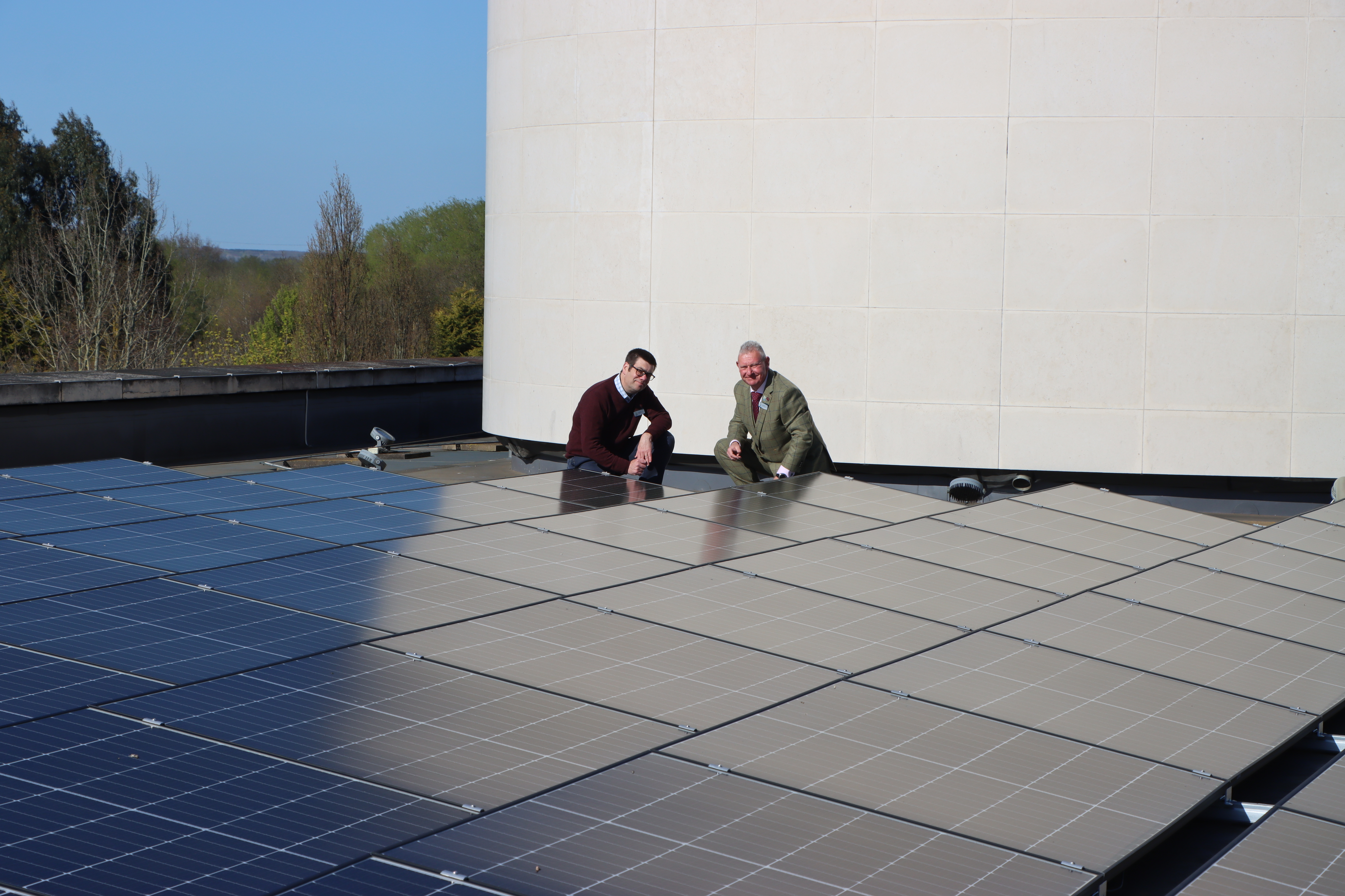 Two men in suits crouch proudly next to a roof full of solar panels.