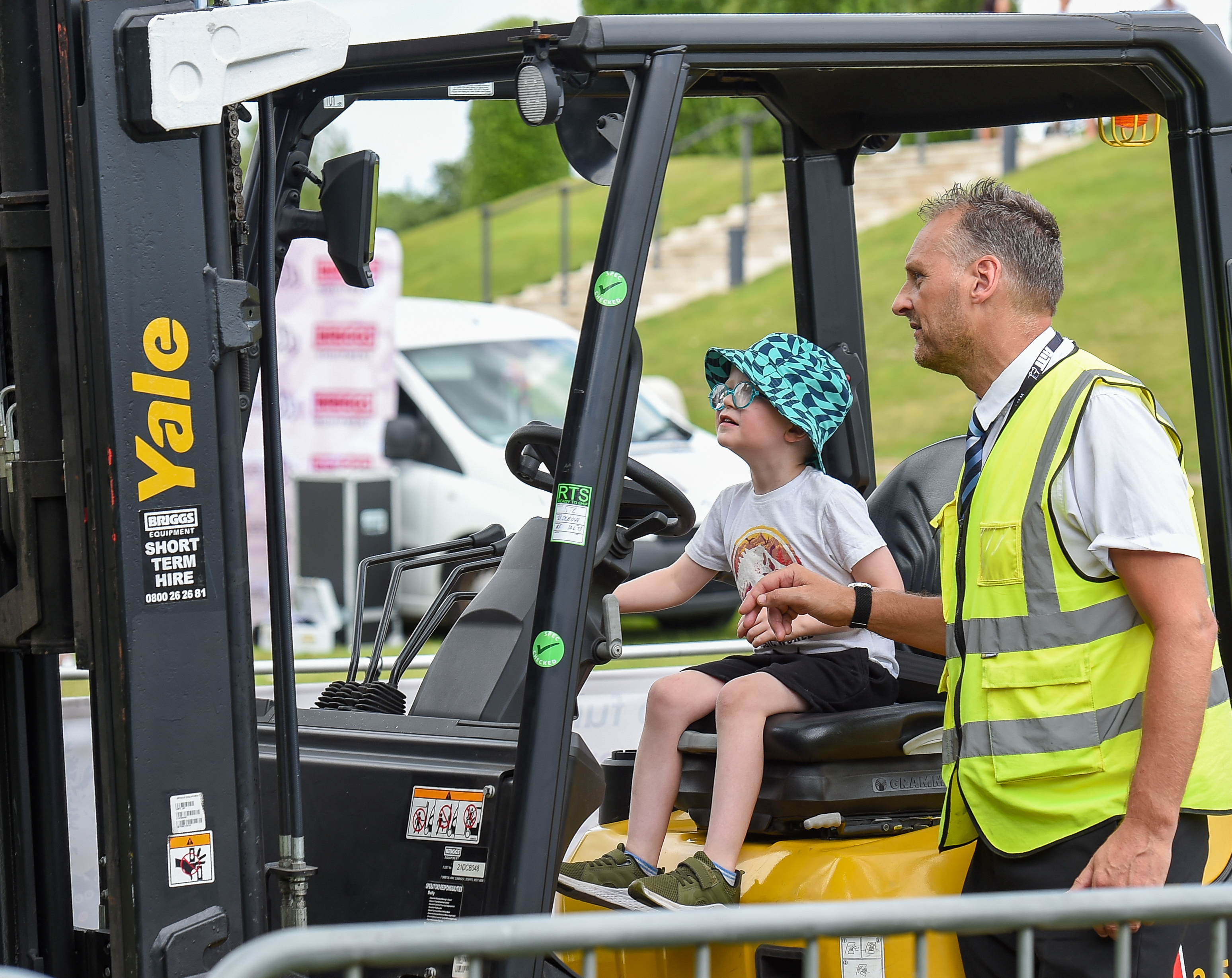A young boy is joined by a briggs equipment employee at the controls of a forklift.