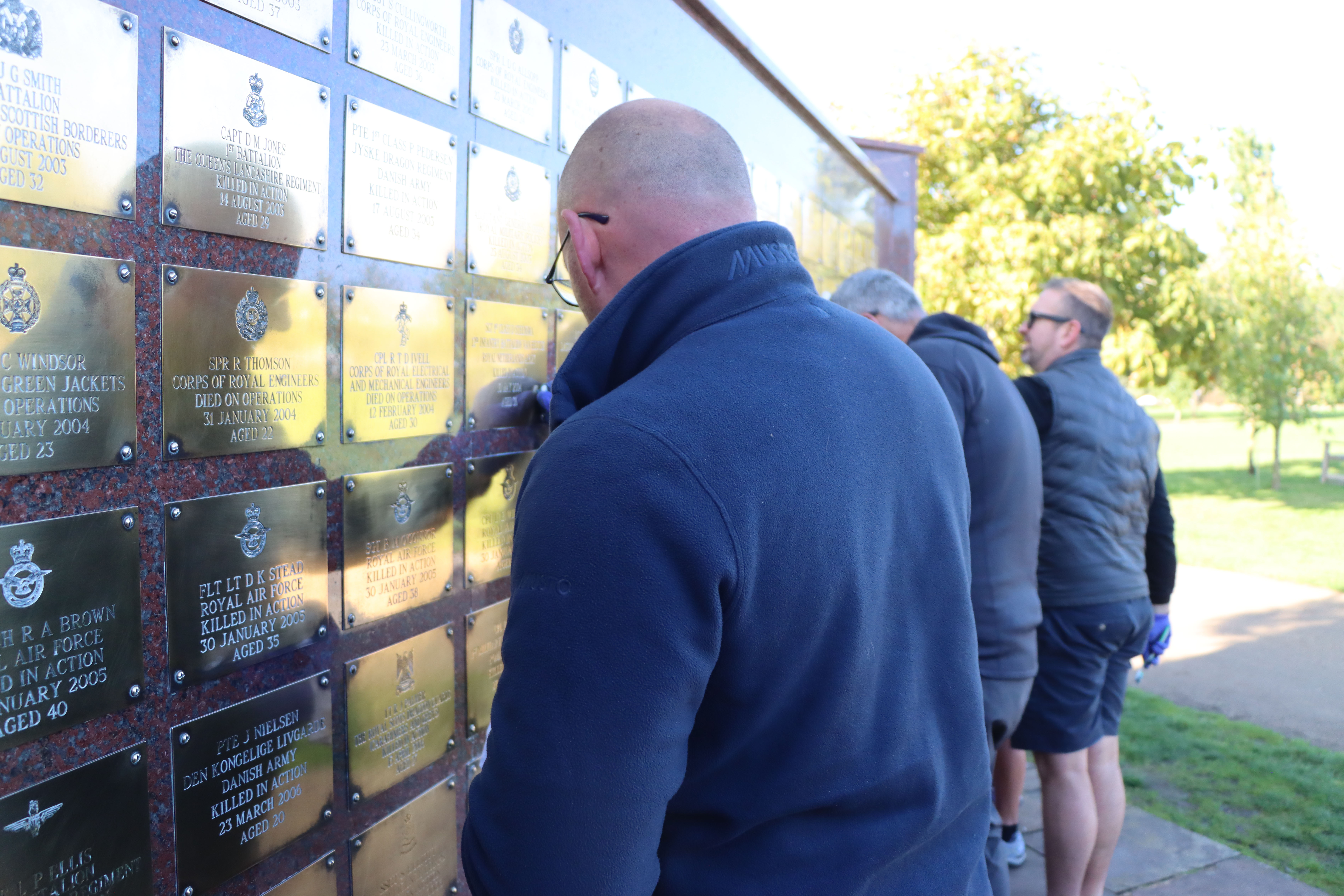 Briggs Equipment employees standing at the Basra Wall memorial, polishing the brass plaques. They are in fleeces as it is a cold, september morning.