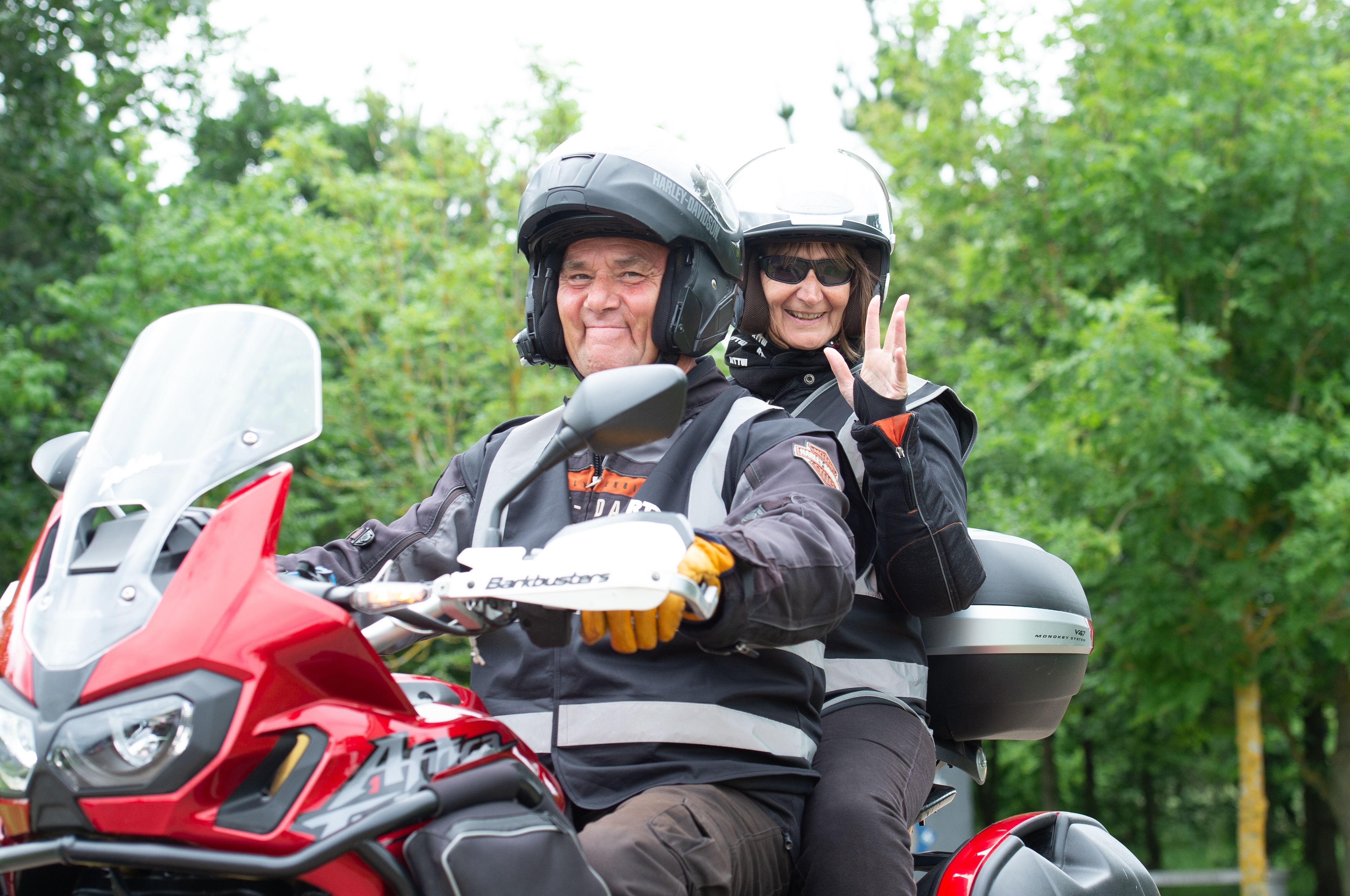A male and female riding on a Motorbike to the National Remembrance Gardens. The lady is waving at the camera, they are both smiling.