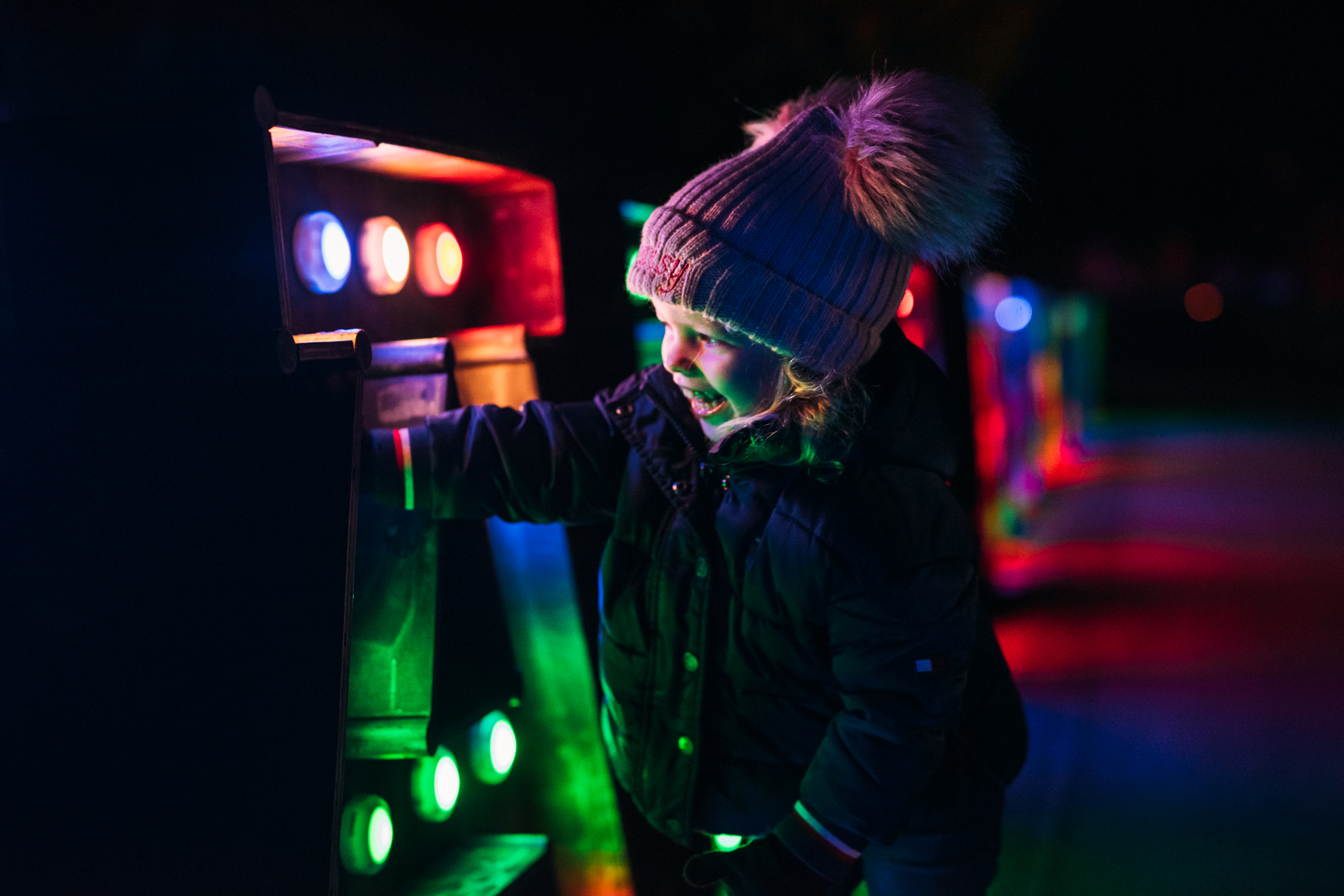 A smiling little girl wearing winter clothing is reaching out and pressing multi-coloured illuminated buttons. The buttons are on large letters that collectively spell out Illuminated.