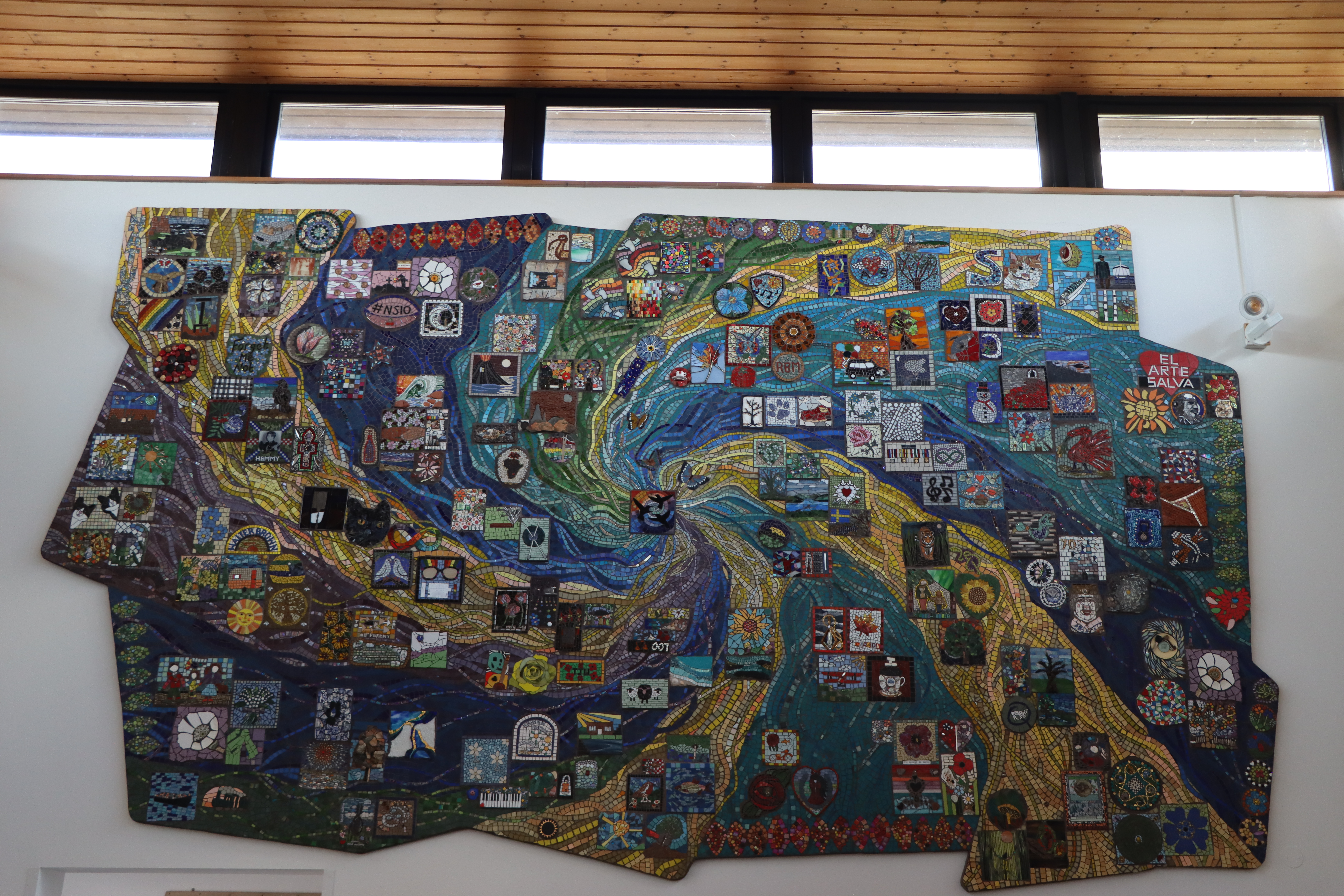 a large mosaic hangs from a white wall. It features a large blue, yellow and green swirl, with individual square mosaics dotted across it.