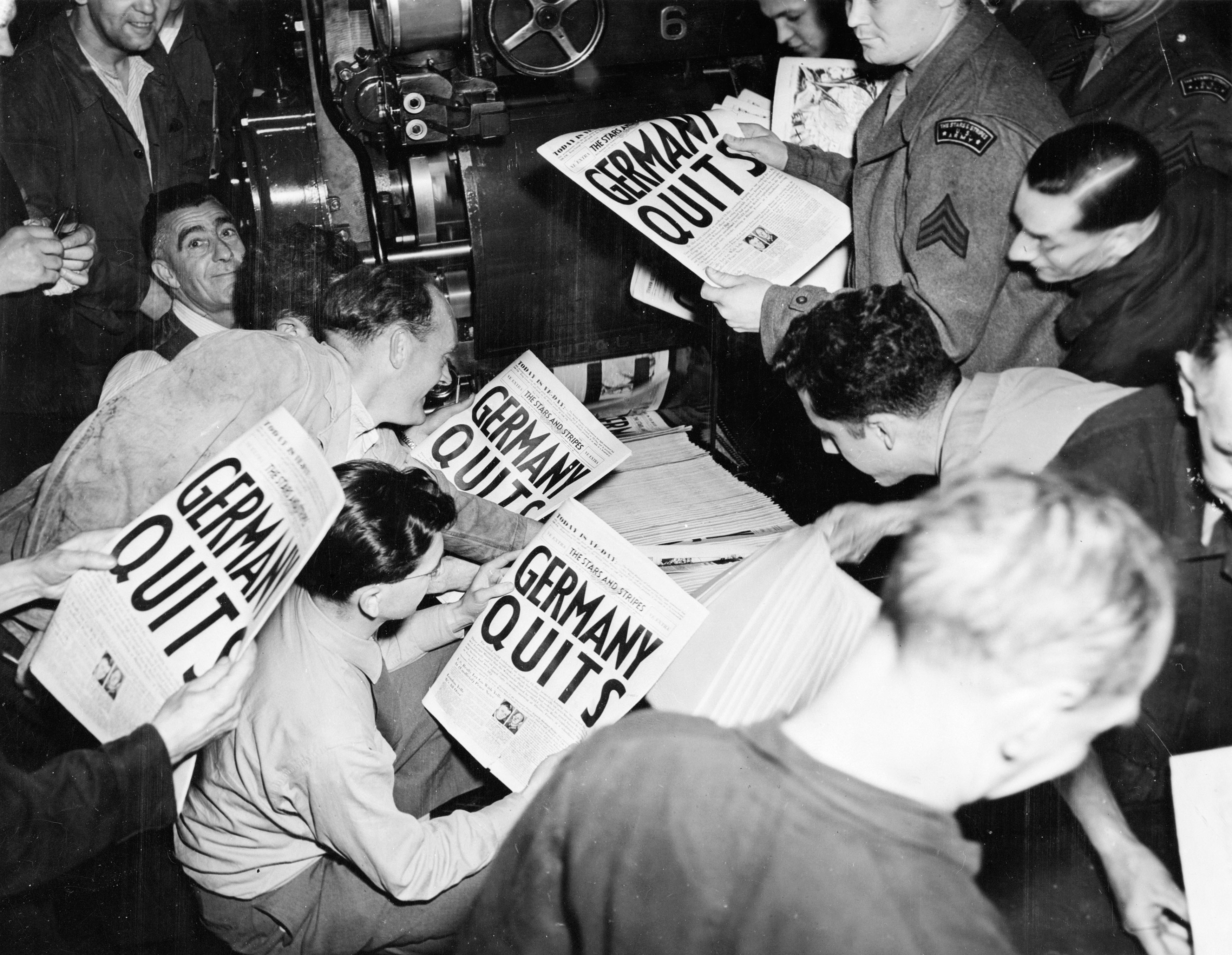 A black and white photograph of eager soldiers pulling copies of the London Times from the press at 9pm on 7 May. Announcing the news of Germany's Surrender, the headline reads GERMANY QUITS.