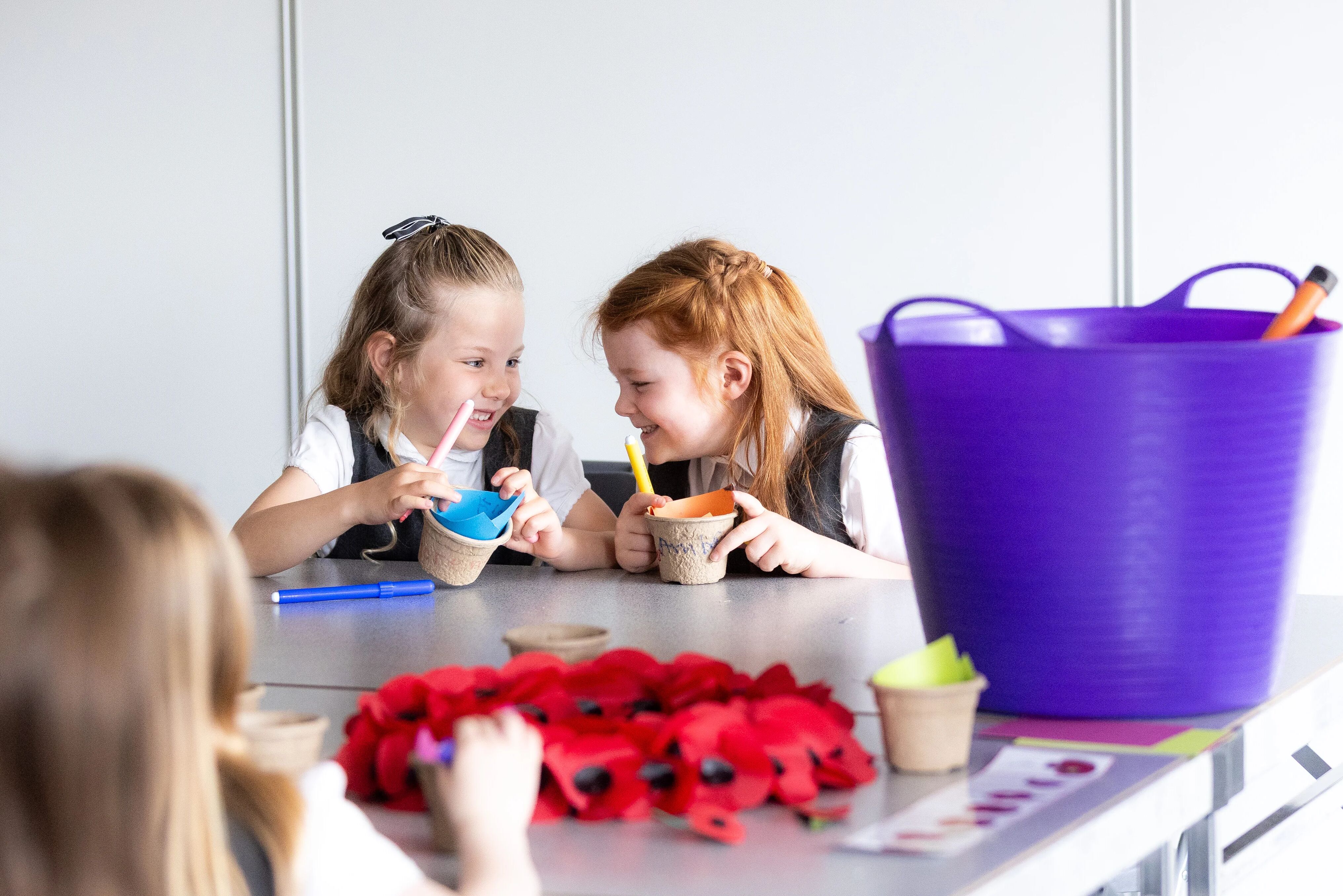 Two girls in key stage one, laughing together while they decorate paper plant pots to plant poppies in as part of an Activity Day.