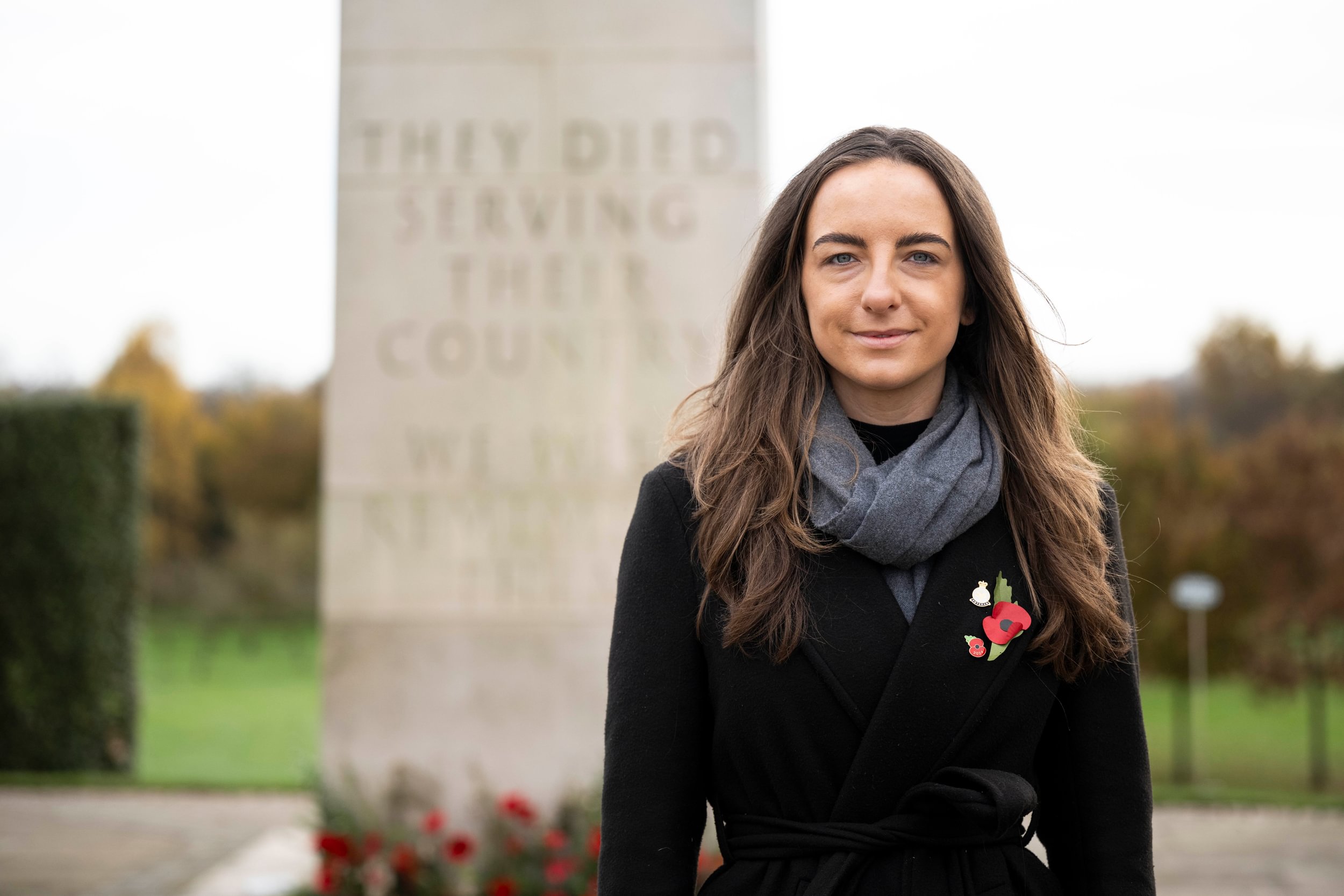 A young woman with long brown hair wears a black coat, with a poppy on its lapel, and a grey scarf. She looks directly at the photographer as her picture is taken.