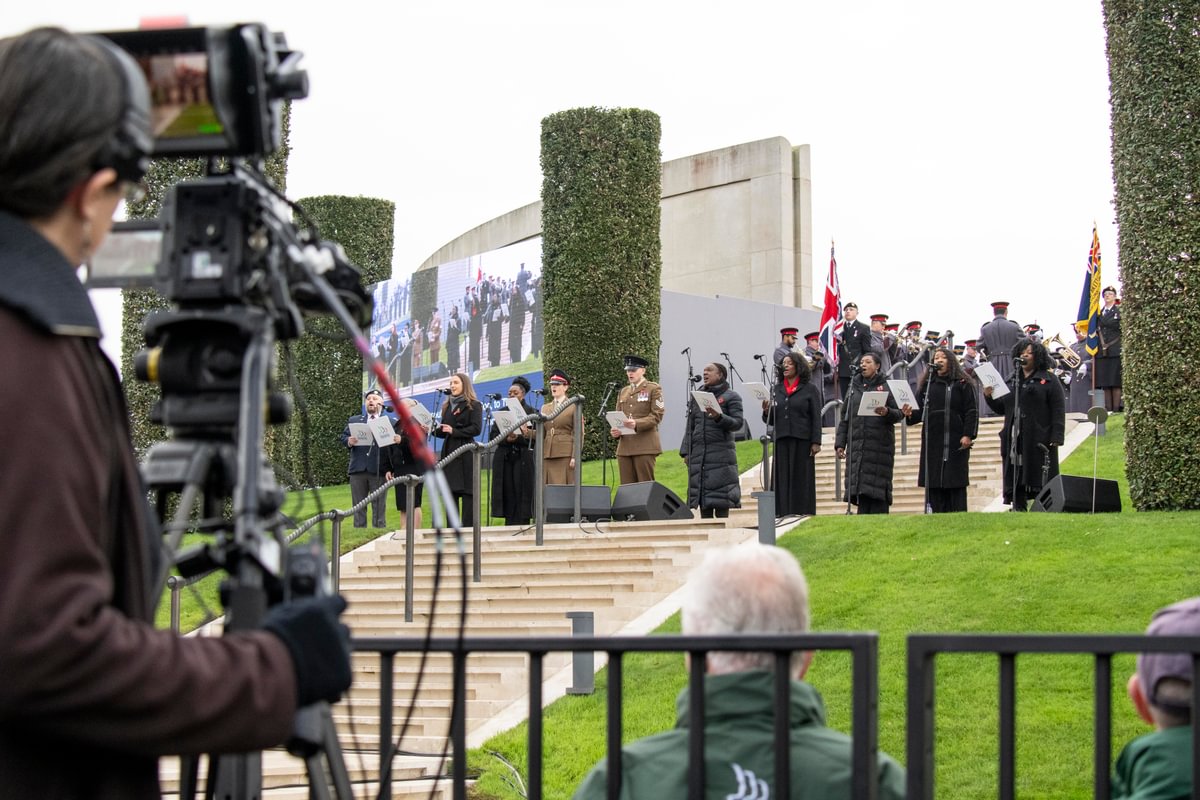 Eleven people stand singing on the Armed Forces Memorial. Behind them at the top of the memorial a military band plays. In the foreground, a person at a camera captures the performance. His footage is shown on a large screen behind the performers.
