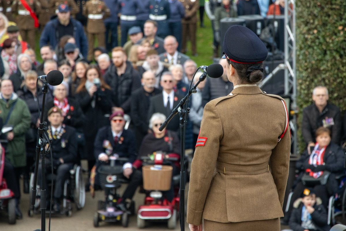 With her back to the photographer a female soldier dressed in a brown formal dress uniform stands in front of a microphone looking down to a crowd below.