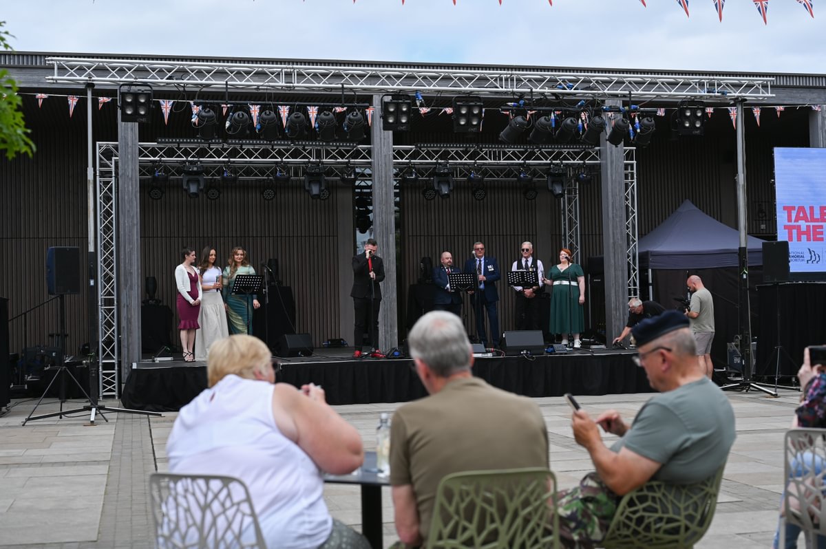 A group of three people sit and watch a stage on which are assembled eight performers singing into microphones