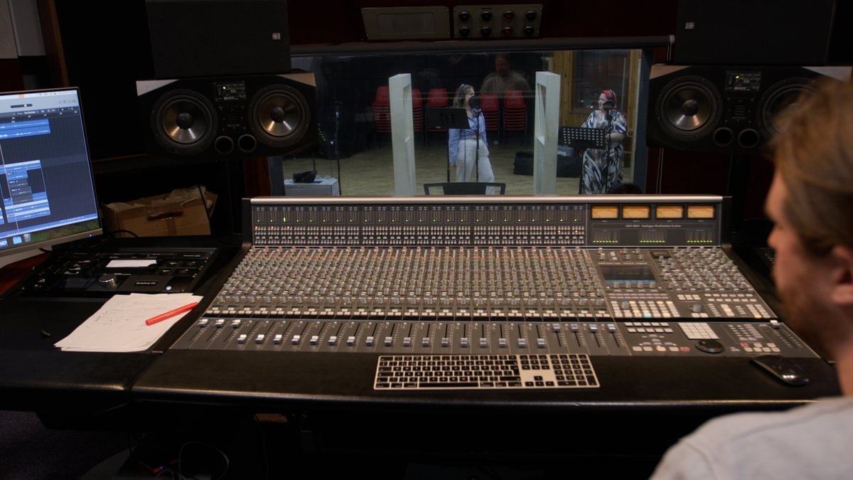 Immediately behind the desk a glass window provides a view into another studio space.  Two women, wearing headphones, stand in this studio ready to record.
