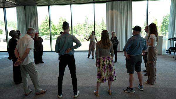 A group of 12 people, most with their backs to the photographer, stand in a semi-circle in a large room. Dressed casually, they all look toward a young woman who is talking.