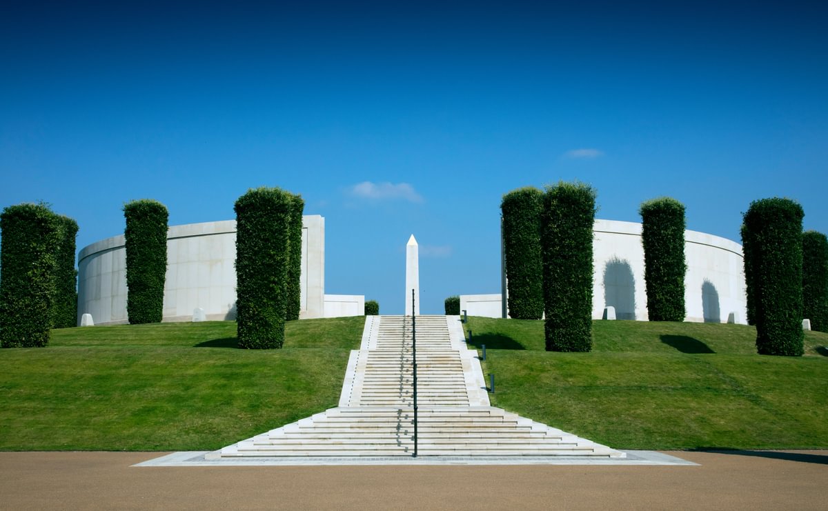 Stairs leading up to curved stone walls surrounded by holme oak trees with a tall stone obelisk in the middle