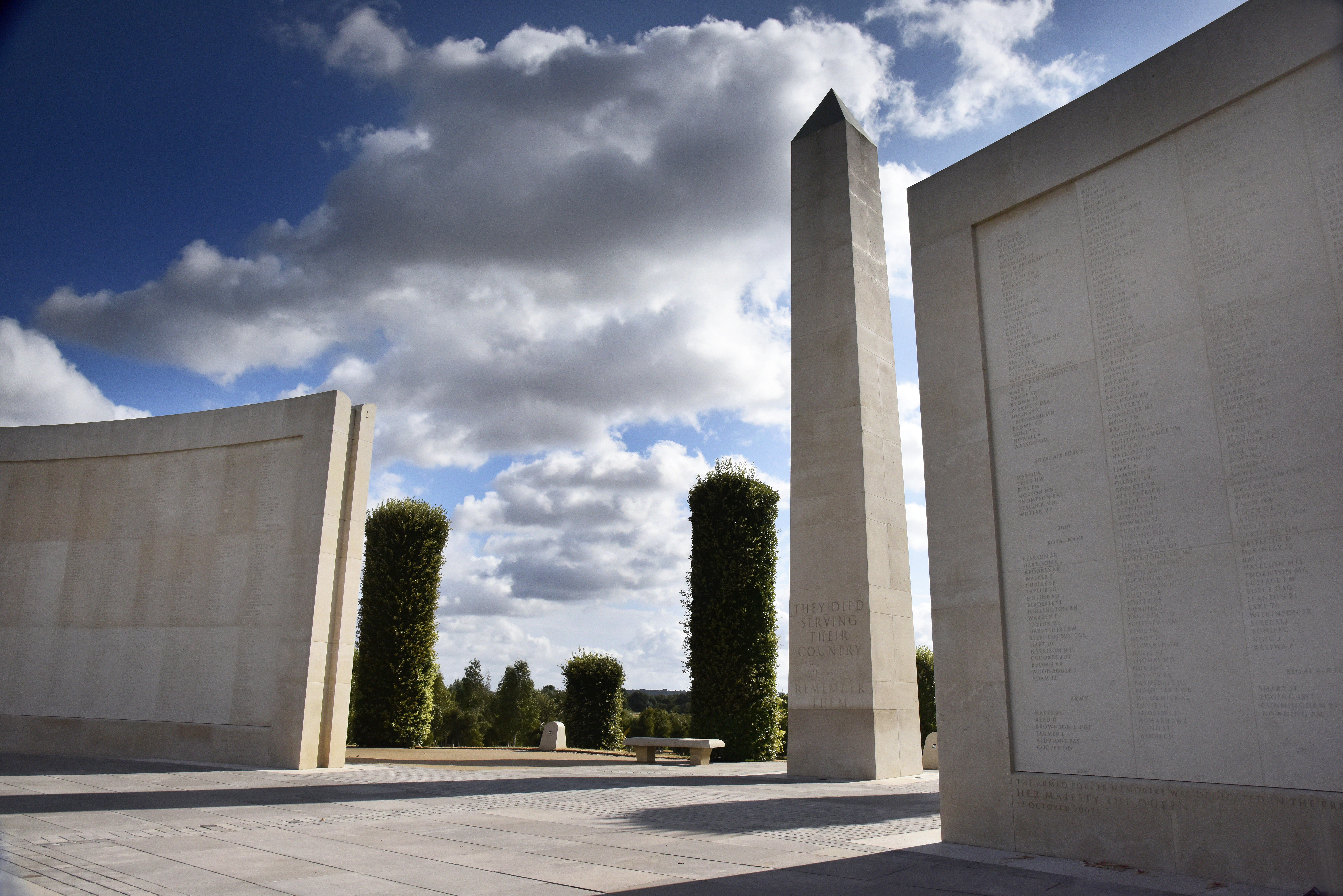 A white stone obelisk stands tall in a gap between two white stone walls, adorned with engraved names of the fallen.