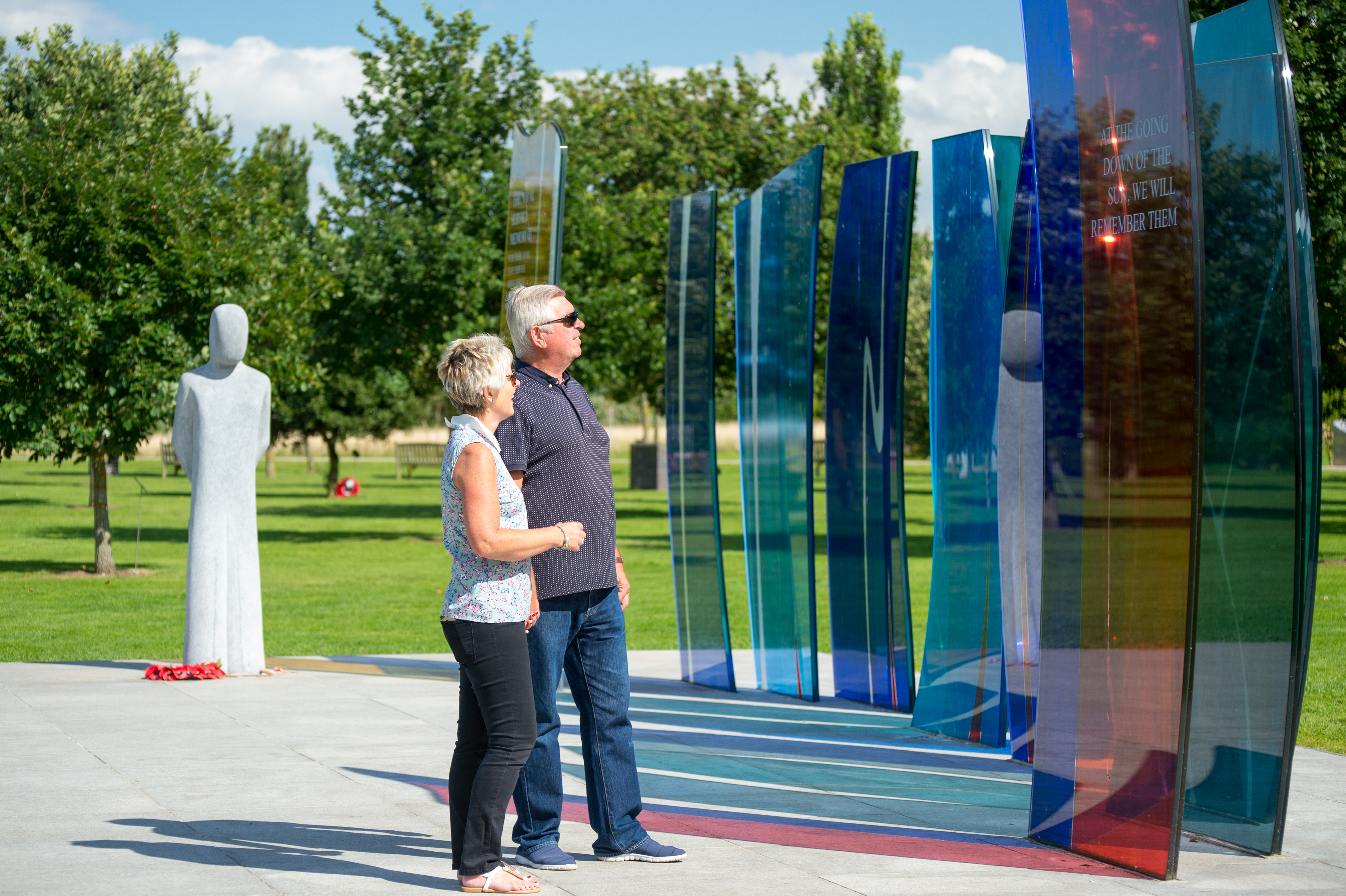 A couple looking up at the colourful glass pains of the Naval Services Memorial. It is a sunny day.