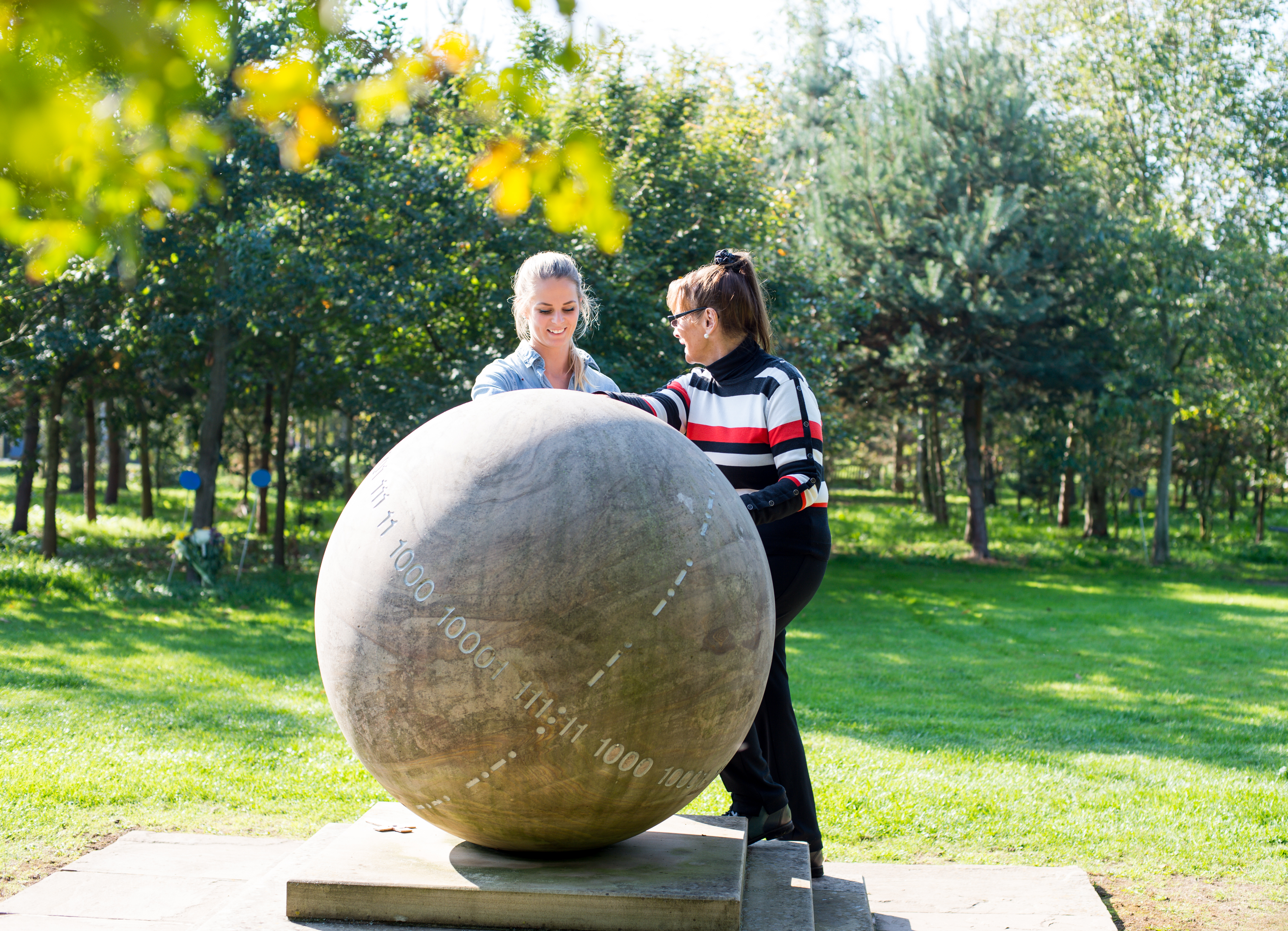 Two visitors look at the GCHQ Memorial. It is a sunny day and they are smiling together.