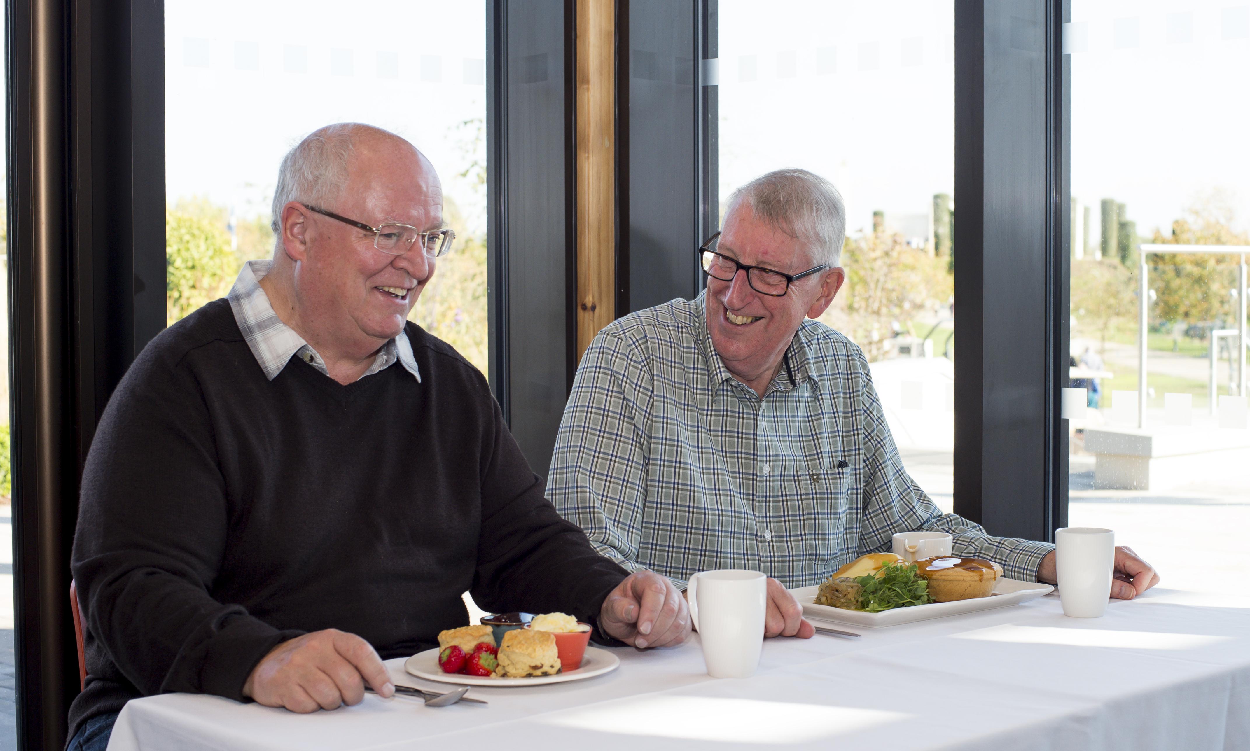 Two male visitors in shirts enjoy a cream tea and pie meal at a clothed table.
