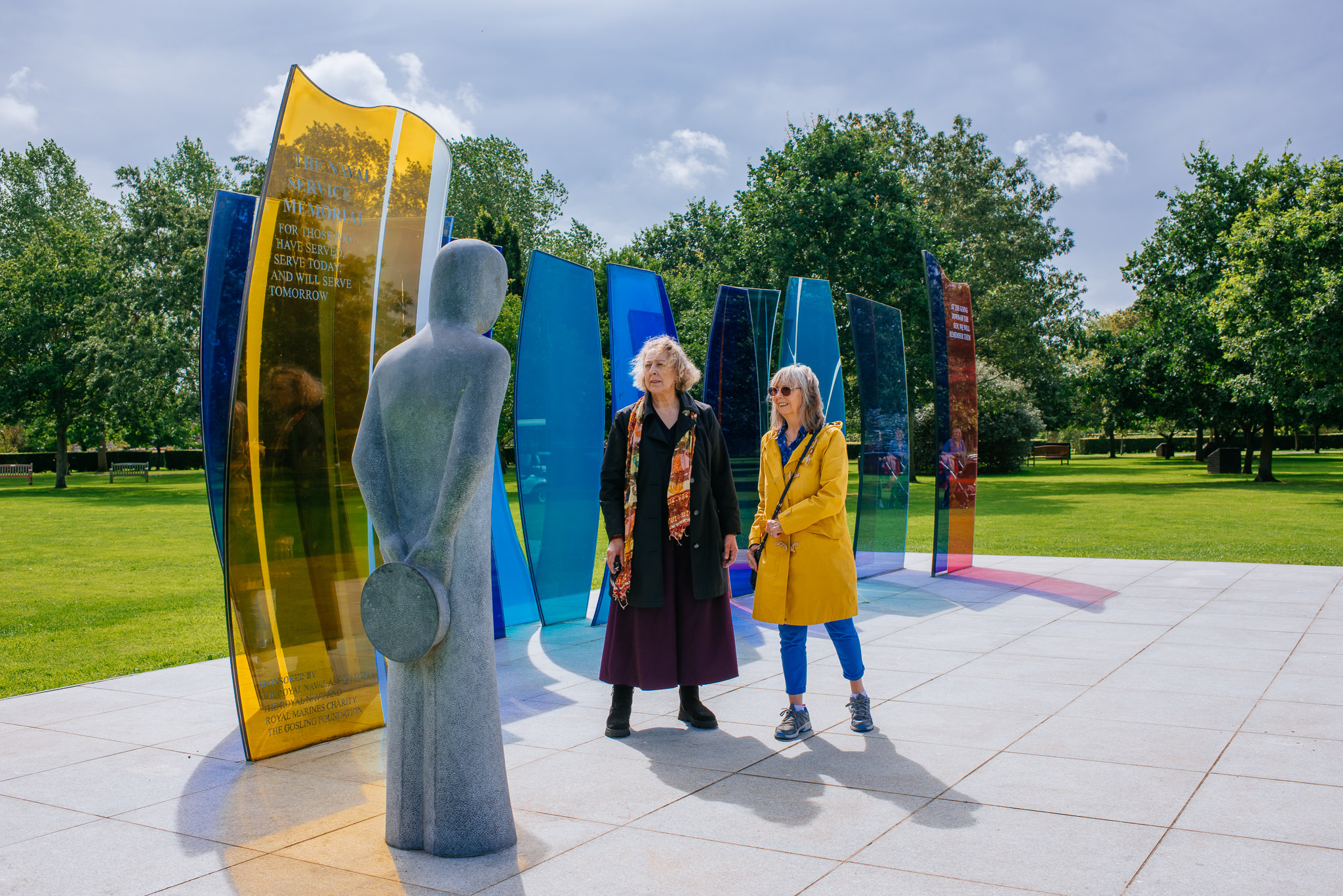 Two female visitors at the Naval Review. They are both in long coats, staring up at a stone sculpture of a sailor.