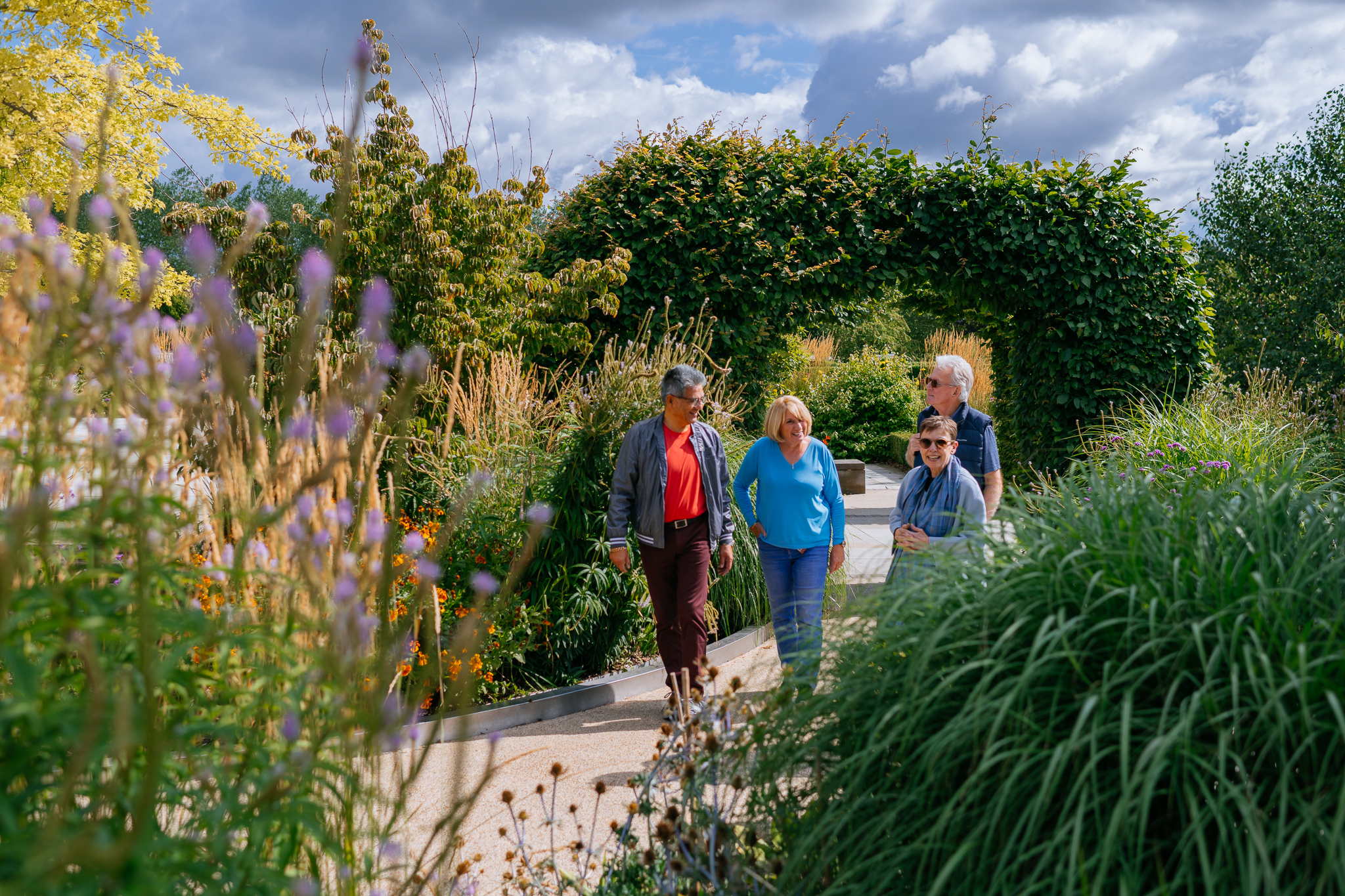 Two women and two men smiling as they walk through the Boyes Garden, surrounded by flowers and greenery. A leafy archway is behind them.