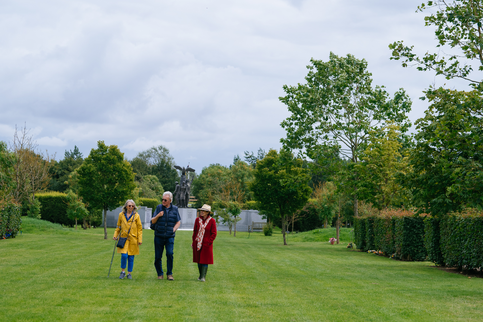 three people  dressed in long coats and scarves walking down a grassed area with a memorial behind them